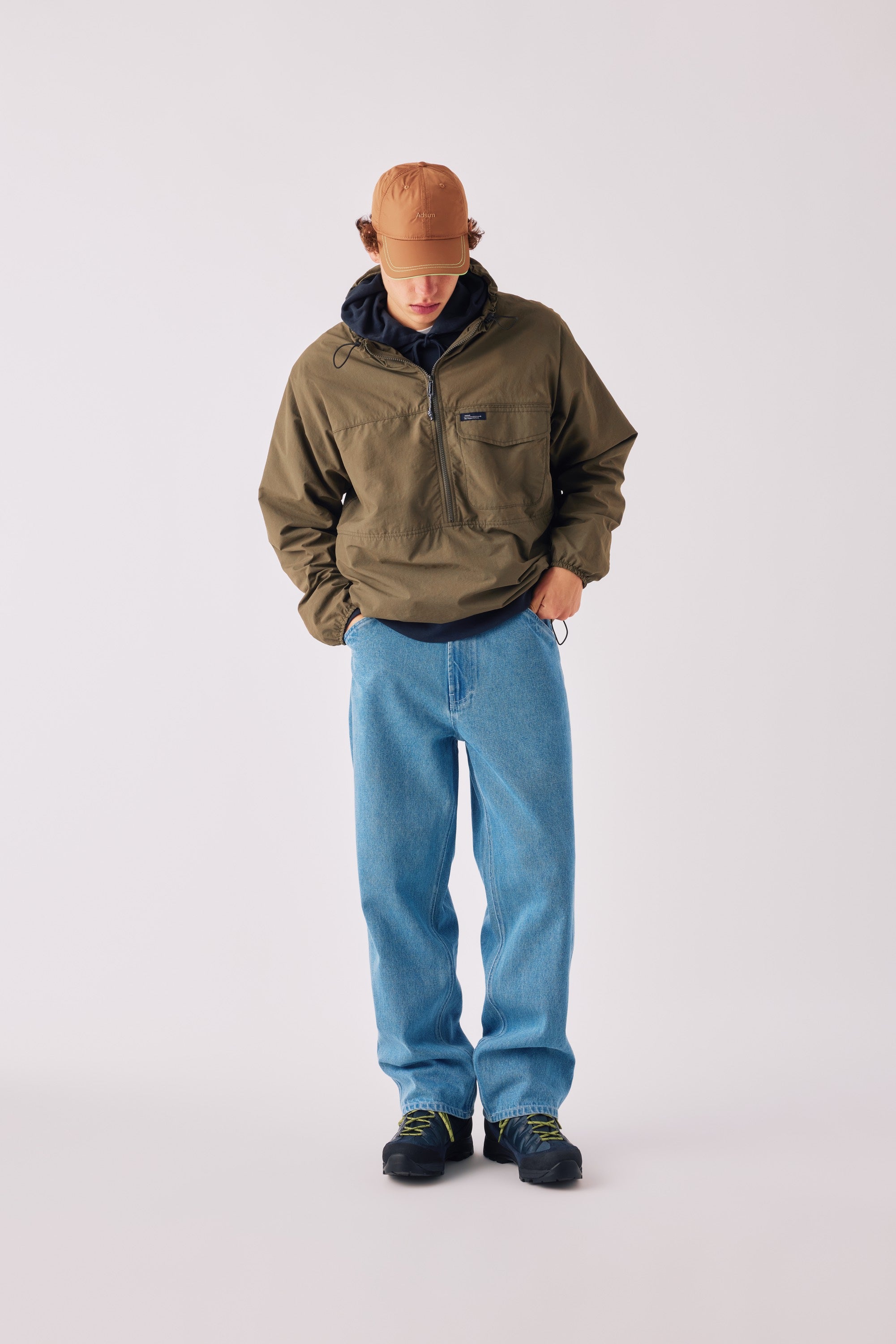 Adsum FW26 — model in an olive half-zip anorak, tan cap and light-wash jeans